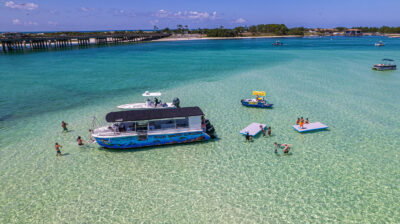 Aerial view of the Crab Island Runner boat with multiple people enjoying themselves in clear blue water in Destin, Florida. The boat has an under water image on the side with the text "Crab Island Runner"