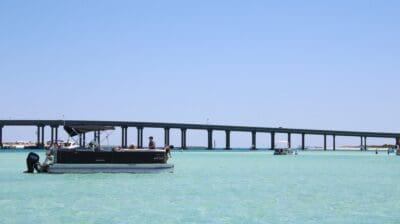 A pontoon boat anchored in clear shallow water at Crab Island with the Destin bridge behind it